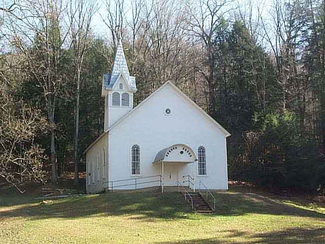 A white woden church with a steeple showing four tiny dormers, and a rain canopy in front, Spruce Grove Church sits on a small hill.