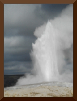 The geyser erupts high in the air against a background of blue sky and fluffy white clouds