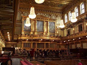 A few musicians in black tuxedos are standing as the orchestra begins to gather for the performance in the heavily gilt ornate music hall