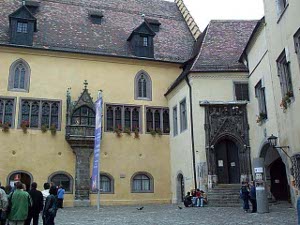 The window on the cream-colored building is surmounted with a cross on a peaked roof and features a stone support down to ground level