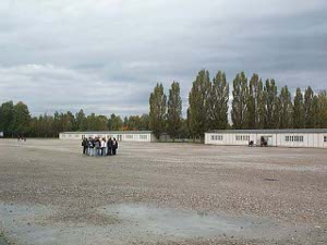 In the distance are two long narrow barracks, with a line of tall trees behind; in the foreground, standing in the middle of the huge yard is a group of about a dozen students of high school age.