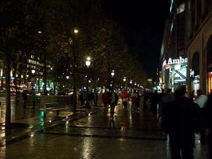 A rainy night, with the lights reflected in the wet pavement, and shoppers and brightly lit stores