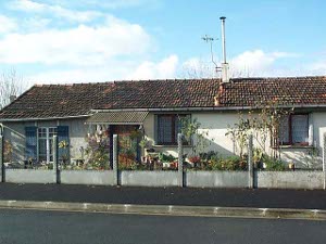 A white stucco ranch house with a tile roof, this suburban home is surrounded by roses and potted plants and flowers