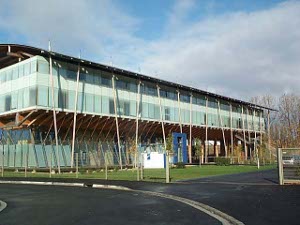 This is a three story building of a modern architecture built of wood the building is painted a pale turquoise with a white horizontal accent line and an overhanging wooden platform above the first story.  Long thin poles serving as columns support this platform and the roof; they extend at varying angles, and the tips of the poles are shaped and painted to resemble pencil tips.