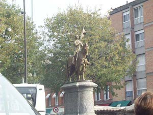 The statue is mounted on a tall granite pedestal; Joan holds a banner and sits astride a horse with the right foreleg raised.  She is looking upward; the whole statue is of a golden color