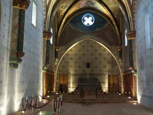 The walls of this church are white with gold crosses, while the arches and nave are decorated tan stone with frescos on the roof; above the altar is a window in the form of a quatrefoil.