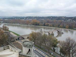 The old bridge consisted of a series of stone arches across the Rhone River.  Some of these arches have now collapsed and the bridge has not been rebuilt, nor has it been torn down.  The river appears brown under a gray sky, curving gently to the left, with bare trees along the banks, and a road on the bank nearest the photographer, who is high on the palace of popes.