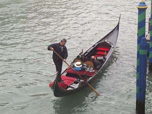 The gondolier is bare-headed, although his straw hat is visible in the voat.  He has the oar engaged with a post on the starboard, far aft, as he uses the leverage to propel the boat and its passenger to the requested destination