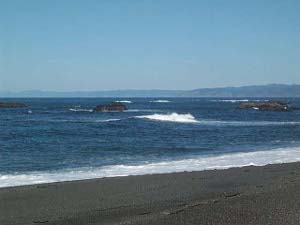 From foreground up: a dark gray sand beach; a ribbon of white sea foam at the shoreline; azure blue Pacific water; dark outcroppings of coastal rocks fringed with white breakers; a distant line of mountains adjoining the coast; and a pale blue winter sky