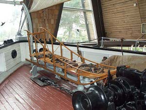 The bow of the St. Roch with a red brown deck and white walls with a black rail, is home to the black anchor windlass and rigging and a long slender oak dogsled
