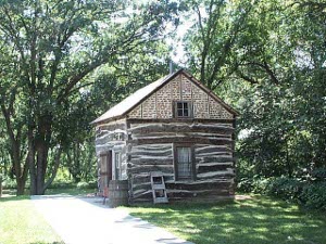 The log cabin is chinked with white plaster and a peaked roof has been added, along with glass windows