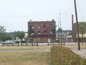 A 3-story apartment building, surrounded by vacant lots, with black iron fire escapes in front, boarded up on the ground floor, windows broken on the upper floors, clearly a center of poverty and crime