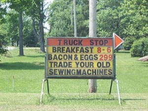 The five line sign reads, TRUCK STOP; BREAKFAST 8-6; BACON & EGGS 299; TRADE YOUR OLD; SEWINGMACHINES