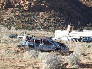 In a sandy desert sits an old junk car; three giant arrows have been crafted appearing to pierce the car.