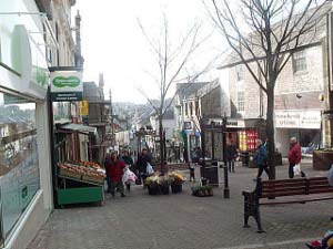 The street is a pedestrian way, on a hill, with trees down the center.  A greengrocer has set out trays of fruit in front of his store, and benches are available in which to rest