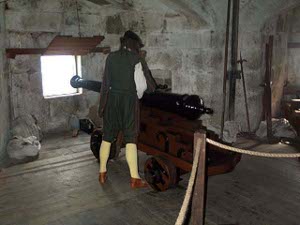A dummy dressed in white shirt, black vest and trousers, knee-length yellow socks and brown shoes stands next to the shiny black cannon on display pointing out a gun port