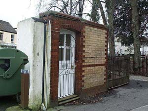 The tiny red and brown brick building, perhaps 5 by 8 feet, has a door but no window.  An iron grating protects the building from theft.  To the left is a green dumpster.