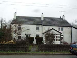 A two story white house with three chimneys stands behind a hedge, against the background of a gray sky