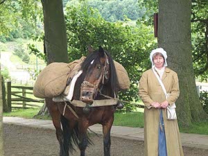 Large canvas sacks are thrown on top of the padded pack saddle, as the horse is led by a woman clad in a long dress, raincoat, and bonnet, and carrying a white bag with food for the horse.