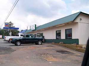 The beige building has a green metal roof, and an overhang, with pickup trucks in front and a Chevron station next door