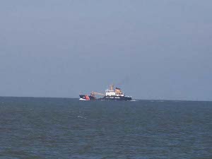 Against the blue green waters and the grey blue sky, the Coast Guard buoy tender in the distance has a black hull, white superstructure, and tan stack.  The cutter is identifiable by the deep dip in the line of the hull where the decks are kept low to allow the heavy buoys to be lifted onto the cutter.
