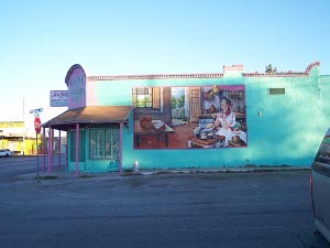 The mural shows a joyous Alice making burritos in a warm brown kitchen painted against the backdrop of a turquoise blue building