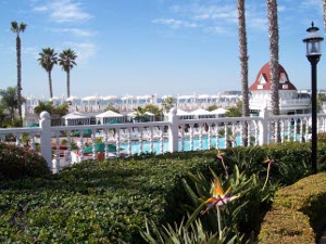 Looking towards the sea, formal gardens are succeeded by an aqua pool surrounded with a white rail fence; on the beach dozens of tables and umbrellas are placed