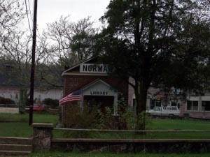Looking like a one-room schoolhouse, the Norman, Arkansas library sits in a green park, with a flagpole planted in front facing a stone wall.