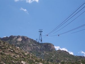 At the base of the mountain, the Sandia Peak Tramway makes a steep ascent over a rocky slope with a bright blue New Mexico sky dotted with white clouds.