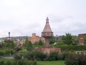 Looking like an inverted cone on a tall pedestal, a reddish brown old fountain is surrounded by a rose garden