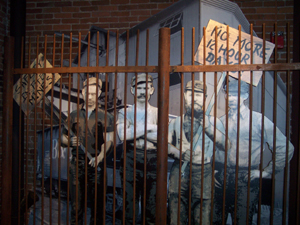 Four striking workers depicted; one holds a sign reading 'No more 12 hour days.'