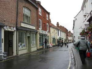 The street, lined with two story buildings (shops below, flats above) narrows noticeably as one approaches the harbor.  The sky is gray.
