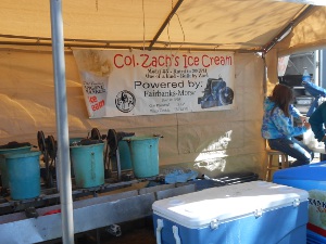 A row of ice cream buckets are agitated by a power belt driven by a steam engine (not shown)