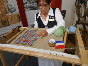 The artist, wearing a dark blue vest over a white blouse and long skirt, sits at the hooking frame, the canvas tightly tensioned as she plants delicate tufts of wool, cut to length, making a pattern that she repeats.