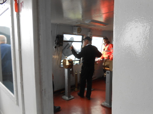 Standing next to the Engine Order Telegraph, and with the Magnetic Binnacle behind him, the skipper is dressed in navy trousers and a white shirt with a navy baseball cap and a long sleeved sweater, giving directions while a crewman in fluorescent safety clothing observes.