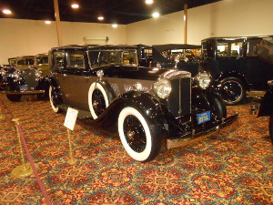 Against a background of similarly colored saloon cars of the same vintage, the Daimler is two-tone black and tope, with gleaming chrome and glass headlamps and hood ornament, and a white-walled spare tire mounted behind the fender.