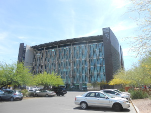 Between two wide grey side walls, the library front shows an attractive diamond pattern of glass and steel framework.  A parking lot with lovely green desert trees is in front.