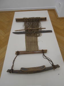 From the top we see a stick holding the warp threads, a narrow stick for the shuttle, athird stick to tap the woof thread down tight, and the sticks holding the woven cloth.  All are simple hand products, shaped into the loom by the weaver herself.