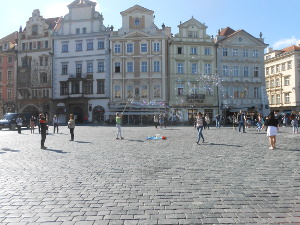 A man dips a net in a large pan of soap suds and waves it in the air, causing bubbles to detach and float across the square in the breeze