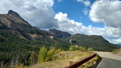 13,000 foot high peaks show a timberline around 11,000 feet; the sky is blue with white clouds, the highway curves around the side of a hill.