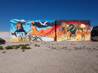 The mural is in two brightly colored panels; on the left a man and a woman ride bicycles; the man is doing a rodeo-style jump, while the womans long skirt stretches out behind her and turns into a brightly colored desert background, where wild desert animals -- javelina, tortoise, and jackalope -- play on their bicycles