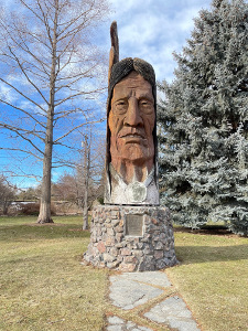 The statue is mounted on a stone pedestal reached by a stone path in the park, and shows a feather rising from the head of the sculpture.  The background is trees against a partly cloudy blue sky