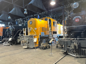Lined up under black exhaust fans are: an orange diesel, a black steam locomotive, a yellow diesel, and Number 93, another steam locomotive.